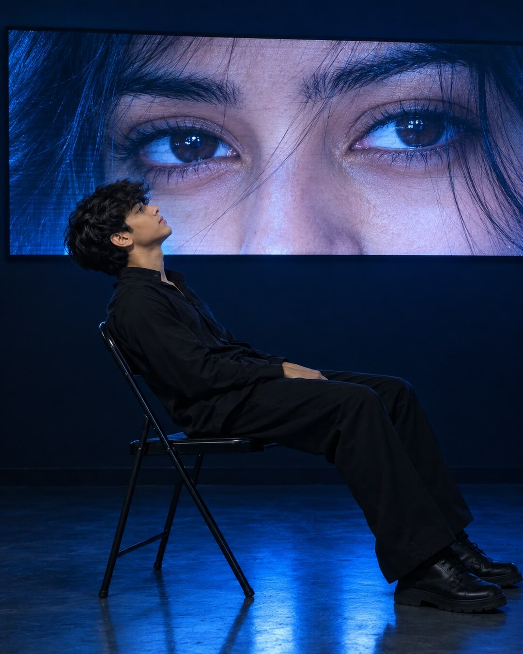 Aesthetic blue neon portrait prompt result - man on chair under giant eyes screen